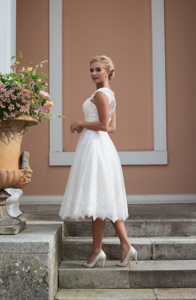 A short ivory lace and tulle gown model is on the steps of a villa