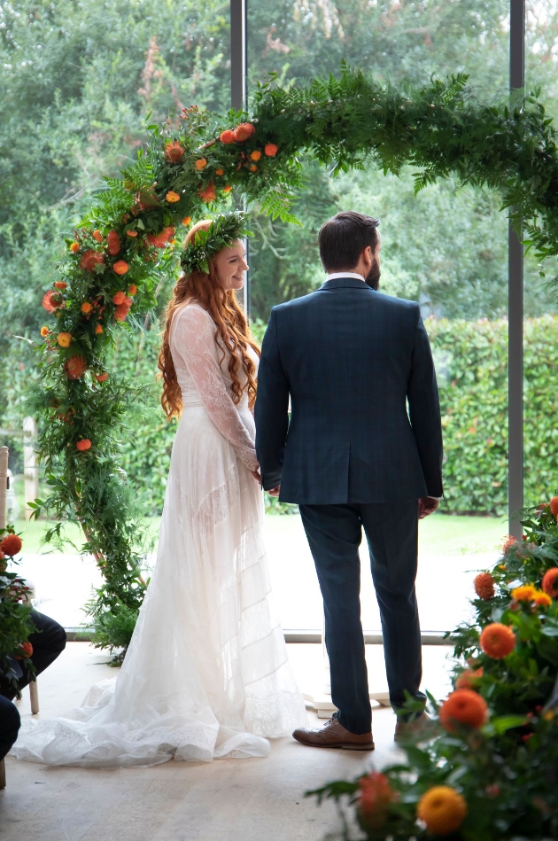 bride and groom in wedding outfits standing in front a moongate floral arch