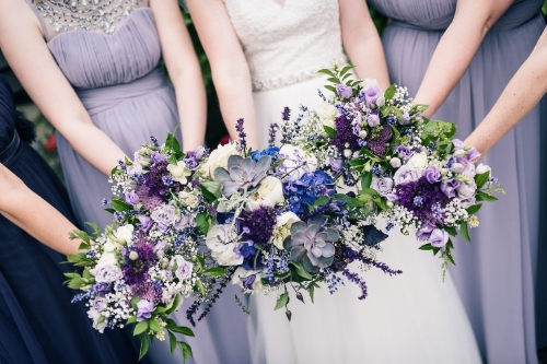 a bride in a white dress, four bridesmaids in purples dresses, everyone is holding bouquets of purple and white flowers