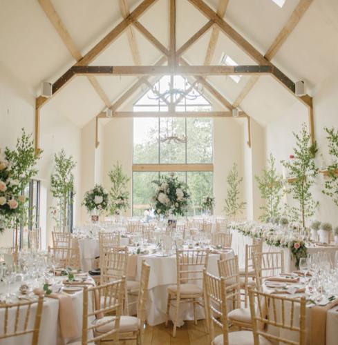 wedding reception room with white walls and wooden beams, tables set up for dinner and feature vases of flowers in centre