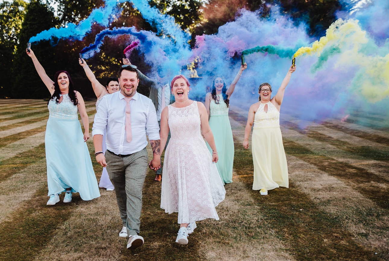 bride and groom in casual wedding gear with their bridesmaids in green dresses all carrying smoke bombs